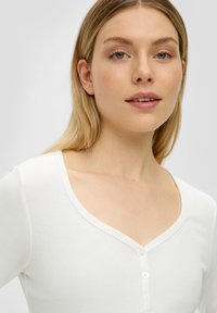 Witte geribde top met een diepe v-hals en een knoopsluiting. De stof lijkt zacht met een gladde textuur. Eenvoudig en veelzijdig ontwerp.