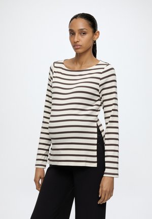 Femme enceinte debout, portant un haut de maternité à manches longues rayé noir et blanc et un pantalon noir, faisant face à un fond uni.