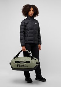 Green duffel bag with black accents and logo, made of durable material. Model wears a black padded jacket and black pants, holding the bag.