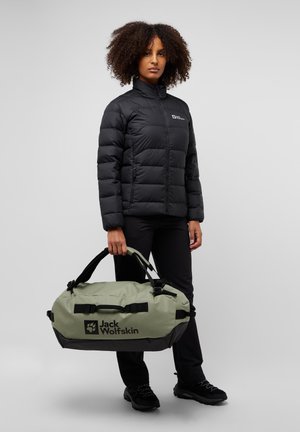 Bolsa de sport verde con acentos negros y logo, hecha de material duradero. El modelo lleva una chaqueta acolchada negra y pantalones negros, sosteniendo la bolsa.