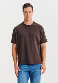 Mężczyzna z krótkimi ciemnymi włosami, ubrany w jednolitą ciemno-brązową koszulkę i niebieskie dżinsy, stoi na tle białego tła.