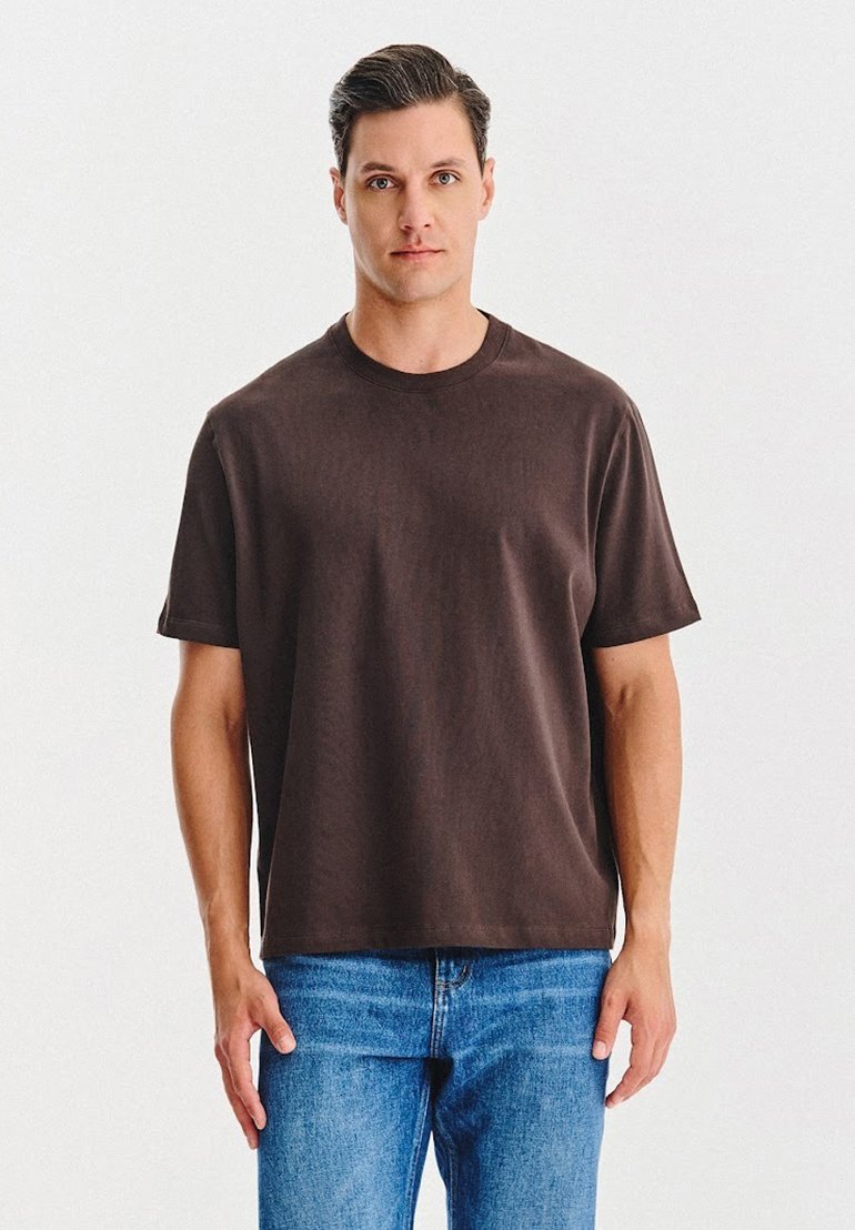 Mężczyzna z krótkimi ciemnymi włosami, ubrany w jednolitą ciemno-brązową koszulkę i niebieskie dżinsy, stoi na tle białego tła.