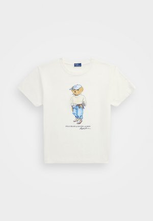 Bela bomba iz bombaža, ki ima podrobno ilustracijo medveda v prostem času, oblečenega v modre kavbojke, belo jopico in modro kapo.