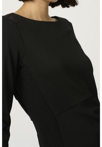 Musta mekko, jossa on tiukka yläosa ja pitkät hihat, joissa on ribbed sivupaneelit ja sileä pinta. Pyöreä pääntie, minimalistinen muotoilu.