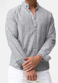 Man draagt een grijs-wit verticaal gestreept overhemd met knopen en een witte broek, handen gekruist voor de taille.