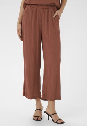 Pantalon ample de couleur rouille avec taille élastique, poches latérales, tissu texturé, porté avec des sandales à talons hauts noires à lanières.