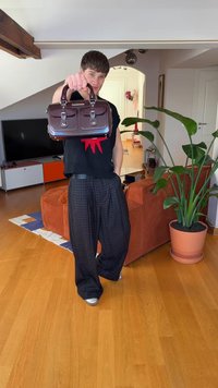 Bruine leren handtas met twee voorvakken en zilverkleurig beslag, vastgehouden door een persoon in een zwarte top en geruite broek, binnenomgeving.