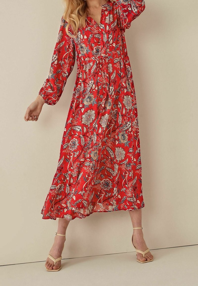 Femme en robe midi fluide rouge à fleurs avec manches longues et talons beiges à brides, debout devant un fond beige uni.