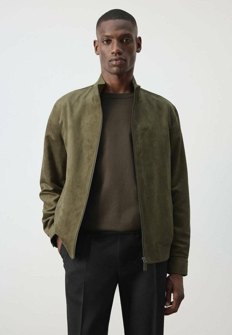 Homme portant une veste en daim vert olive sur un pull vert foncé et un pantalon noir, debout les mains dans les poches devant un fond uni.