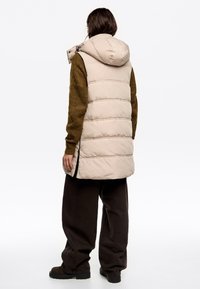 Gilet imbottito beige con cappuccio, caratterizzato da cuciture verticali, zip laterali e una vestibilità rilassata, abbinato a un maglione e pantaloni marrone scuro.