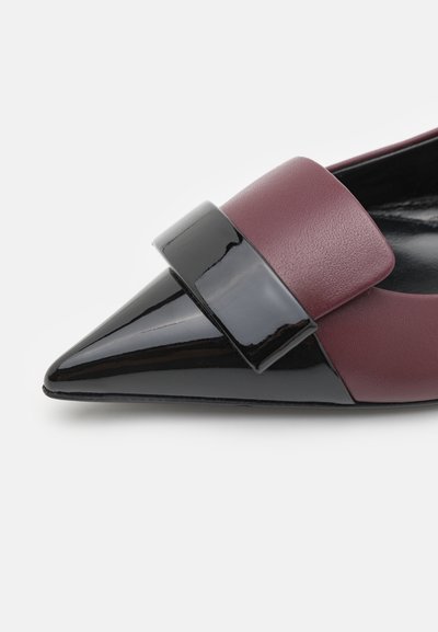 Zapato de charol burdeos con punta afilada y un lazo negro brillante, presenta una textura suave y acabados contrastantes.