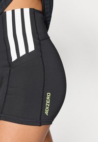 Černé šortky s vysokým pasem, se třemi bílými svislými pruhy na přední straně a limetkově zeleným logem "ADIZERO" na boku, vyrobené z elastického materiálu.