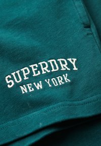Türkiser Baumwollstoff mit gesticktem "SUPERDRY NEW YORK"-Logo in Weiß. Einfaches Design und glatte Textur ohne zusätzliche Muster.