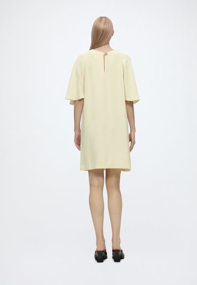 Femme aux longs cheveux blonds portant une robe ample jaune pâle jusqu'aux genoux et des chaussures noires à petits talons, debout de dos sur un fond blanc.