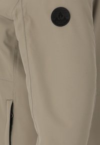 Beige mjukskaljacka med slät struktur, med en svart rund logotyp på överarmen och kontrasterande sömnadsdetaljer.