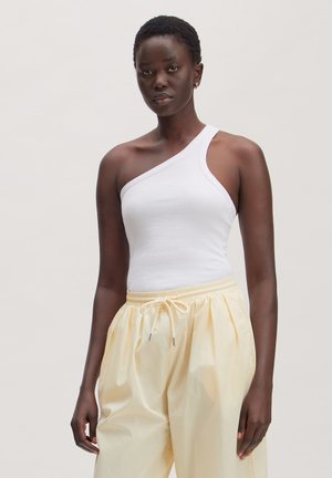 Vrouw met kort krullend haar, die een witte top met één schouder draagt en losse lichtgele broek, staand tegen een effen achtergrond.