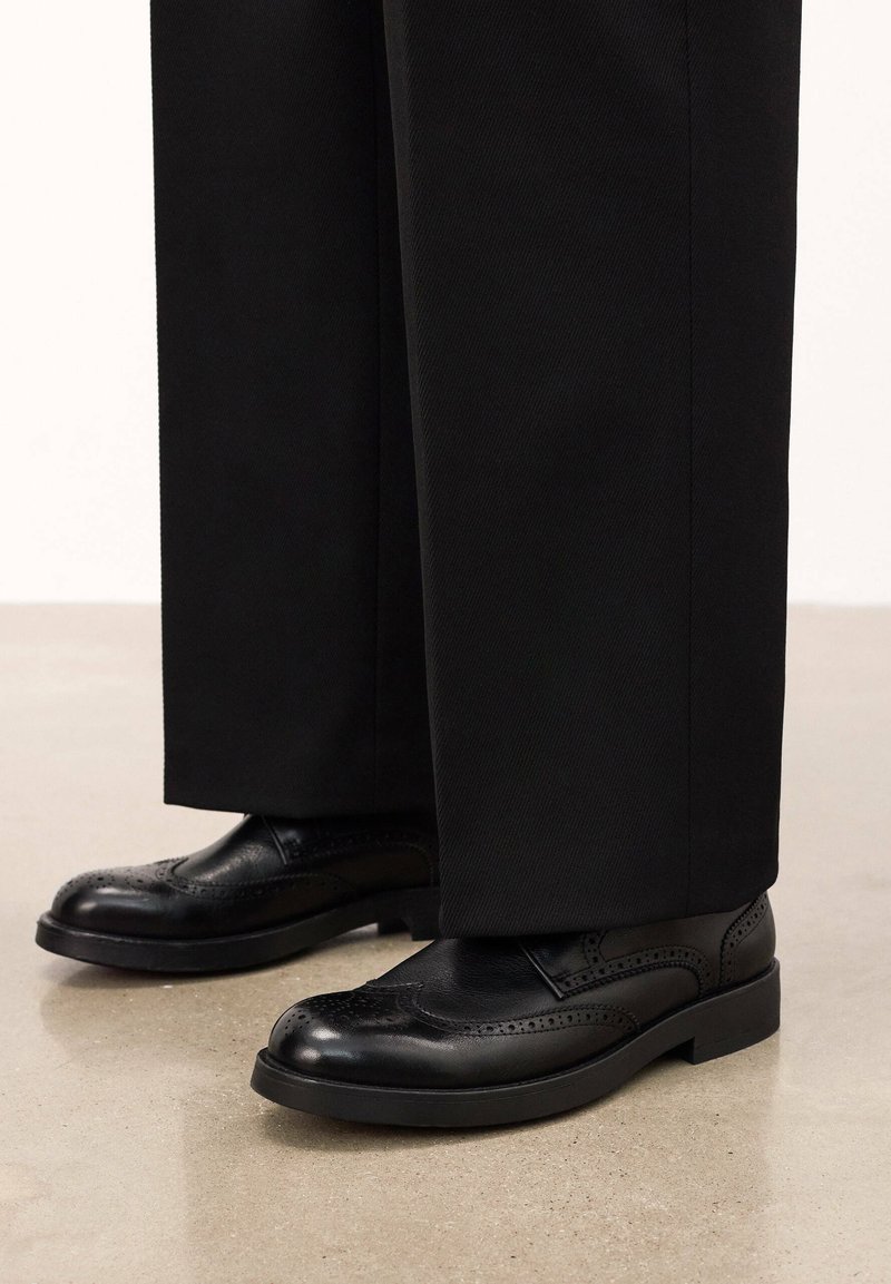 Pantofi de piele negri cu detalii perforate, vârf rotund și talpă plată, asortați cu pantaloni negri. Textură netedă, finisaj lustruit.
