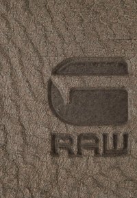 Braunes strukturiertes Leder mit geprägten Buchstaben "G" und "RAW", sichtbaren Rissen und einem robusten Erscheinungsbild.