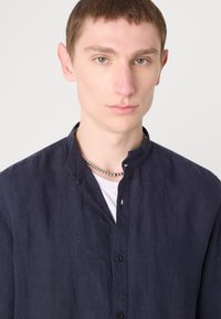 Mørkeblå skjorte med knapper og stående krage, laget av et teksturert stoff. Bært over en hvit T-skjorte, har en subtil placket-detalj.
