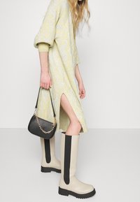 Knähöga beiga gummistövlar med svarta randdetaljer, tillsammans med en texturerad gul klänning med vågmönster och en sidoslits. Svart handväska.