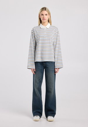 Femme blonde portant un polo à manches longues rayé blanc et marron, jean bleu foncé à jambes larges et baskets blanches, debout devant un fond uni.