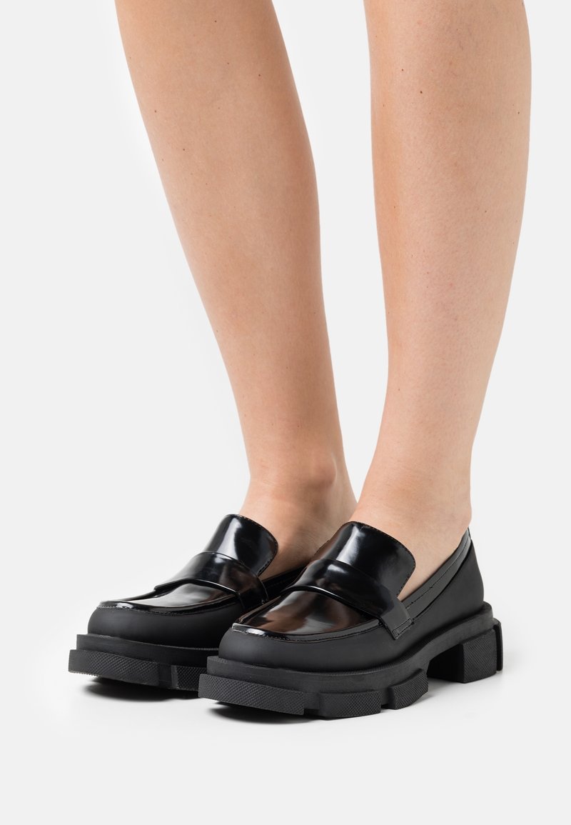 Jambes portant des mocassins noirs à semelles épaisses et dessus en cuir brillant, debout sur un fond blanc.