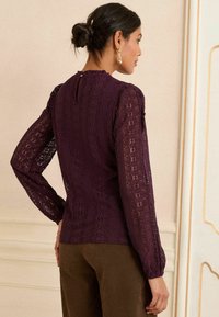 Violetti pitsilöysi pitkähihaisella, tekstuuripinnalla, korkealla kauluksella ja takana ohuella avauksella, joka on yhdistetty ruskeisiin housuihin.