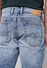 Vue de dos d'un jean en denim bleu clair porté par une personne avec un t-shirt noir, avec deux poches arrière et une étiquette de marque en cuir marron.