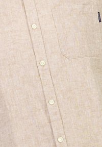 Camicia in lino beige chiaro con chiusura anteriore a bottoni, taschino sul petto e una trama sottile. Quattro bottoni visibili lungo la patta.