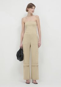 Beige trägerloser Jumpsuit mit weitem Bein, Fronttaschen und einer glatten Textur. Das Model hält eine schwarze Tasche und trägt hochhackige Sandalen.