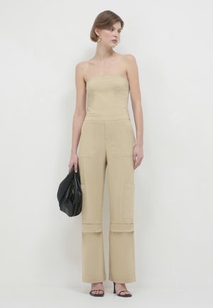 Beige trägerloser Jumpsuit mit weitem Bein, Fronttaschen und einer glatten Textur. Das Model hält eine schwarze Tasche und trägt hochhackige Sandalen.