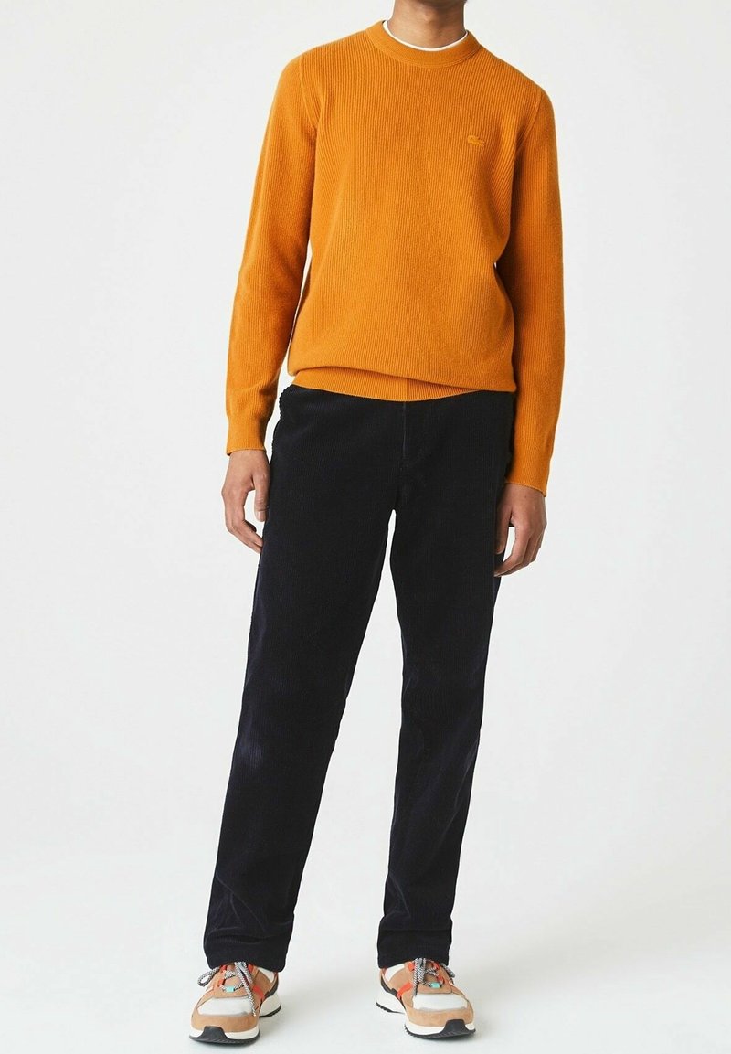 Personne portant un pull orange à manches longues, un pantalon noir et des baskets beiges avec des détails orange et blanc, debout devant un fond blanc uni.