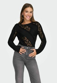 Black long-sleeve top featuring lace fabric with a textured, diagonal design. Paired with high-waisted gray denim jeans.