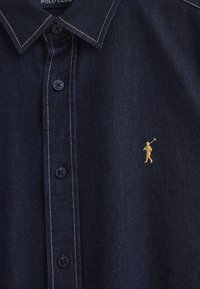 Mörkblå denimskjorta med en strukturerad krage, gyllene sömmar och fem svarta knappar. Har en liten gyllene logotyp av en golfare på vänster sida.