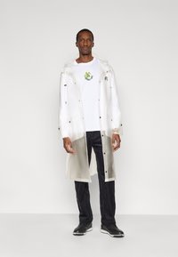 Διαφανές αδιάβροχο μήκους έως το γόνατο με κουμπιά πατιλέτα, ρυθμιζόμενη κουκούλα, φορεμένο πάνω από λευκό πουκάμισο με πράσινο γραφικό. Συνδυασμένο με σκούρο, ριγωτό παντελόνι.