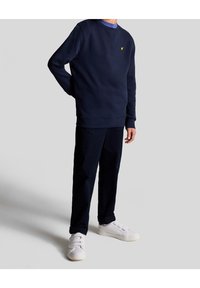 Felpa navy con collo a giro e piccolo logo giallo, abbinata a pantaloni navy e scarpe bianche con design a strappo in velcro.