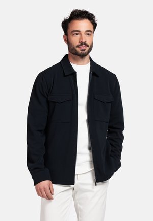 Black, zip-up jacket with two chest pockets, made of soft fabric. Features a collared neck and is worn over a white shirt.