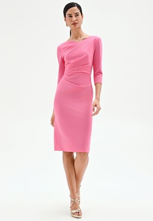 Femme en robe rose mi-longue à manches trois-quarts, debout les jambes croisées et portant des talons dorés à brides.