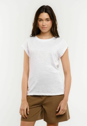 Tânără cu păr lung și închis la culoare, în picioare, purtând un tricou alb simplu cu mâneci scurte și pantaloni scurți maro până la genunchi, pe un fundal simplu.