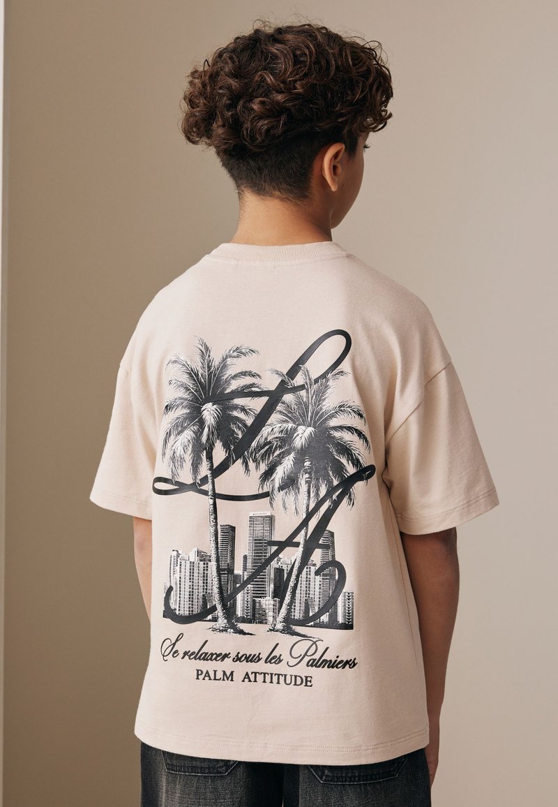 Boy with curly hair wearing a beige t-shirt with palm trees, cityscape, and text "Se relaxer sous les Palmiers Palm Attitude" on the back.