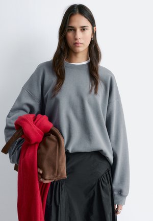 Kvinde med langt mørkt hår iført grå sweatshirt og sort nederdel, holder en brun taske og et rødt tørklæde mod en lys baggrund.