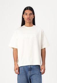 Kremfarget oversized bomulls t-skjorte med korte ermer, med preget "GUESS"-logo på forsiden, kombinert med blå denim jeans.