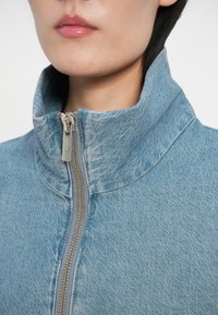 Světle modrá prošívaná bunda se stojáčkem, vyrobená z materiálu podobného džínovině. Má viditelný zip a jemnou strukturu.