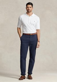 Mand iført en hvid poloshirt, marineblå bukser, brun bælte, brune sko og et ur, stående med den ene hånd i lommen.