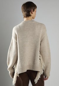 Osoba w oversize'owym beżowym swetrze dzianinowym z frędzlami po bokach, zestawiona z ciemnobrązowymi spodniami, pokazana od tyłu na szarym tle.