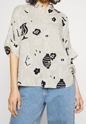 Femme portant une chemise beige ample à boutons avec des motifs floraux et de visages abstraits en noir et blanc, associée à un jean bleu clair.