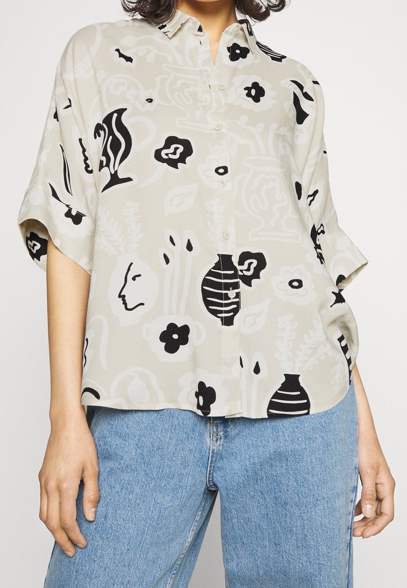 Femme portant une chemise beige ample à boutons avec des motifs floraux et de visages abstraits en noir et blanc, associée à un jean bleu clair.