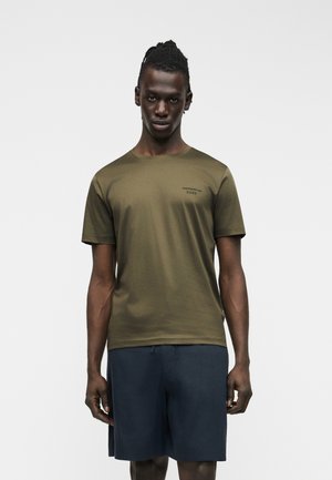 Άνδρας με ντραντλοκς που φοράει κοντομάνικο πουκάμισο ελιάς και σορτς ναυτικού μπλε, στέκεται μπροστά σε ένα απλό ανοιχτό φόντο.