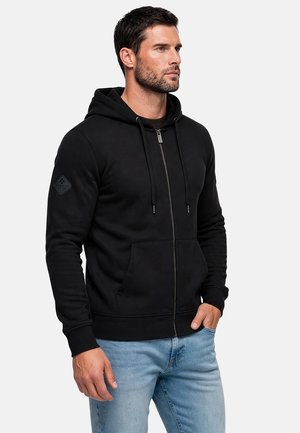 Uomo che indossa una felpa nera con zip e cappuccio, con tasca frontale, abbinata a jeans azzurri chiari, in piedi davanti a uno sfondo semplice.