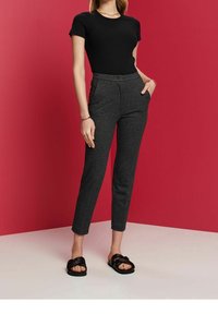 Žena v černém tričku s krátkým rukávem, šedých texturovaných kalhotách po kotníky, černých sandálech a zlatých špercích stojí na červeném pozadí.